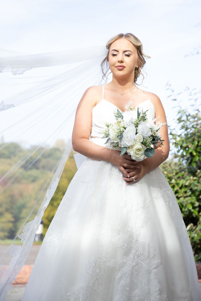 Beautiful bride in white ballgown wedding dress holds elegant bouquet of white roses and greenery against misty backdrop.
