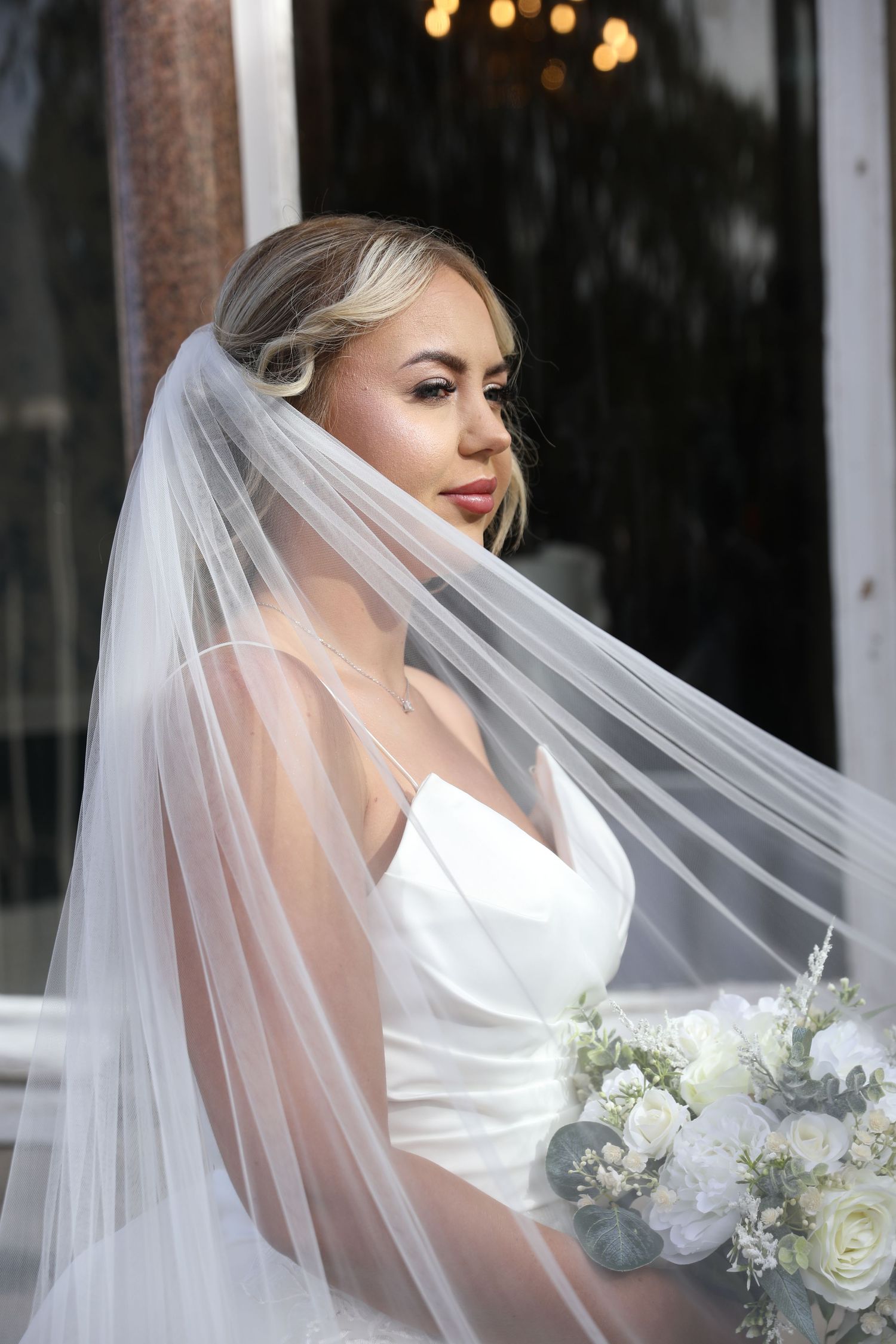 A bride in a white wedding dress and flowing veil poses with a white rose bouquet against a dark background.