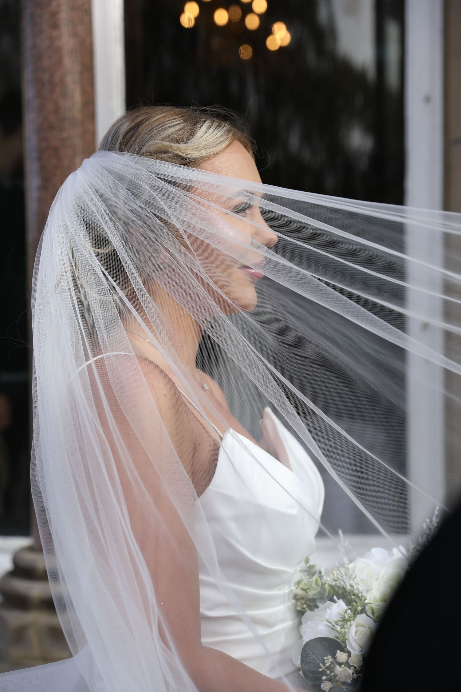 A bride in a white wedding dress and flowing veil holds a white bouquet against a dark background.