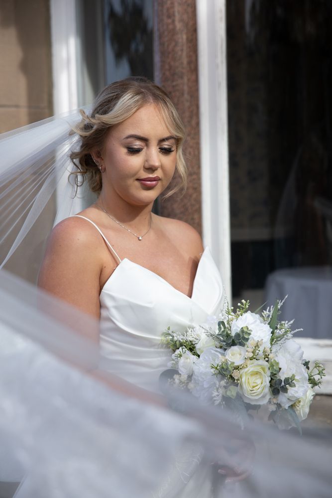A bride in a white dress holds a delicate bouquet of white roses and greenery while looking down.