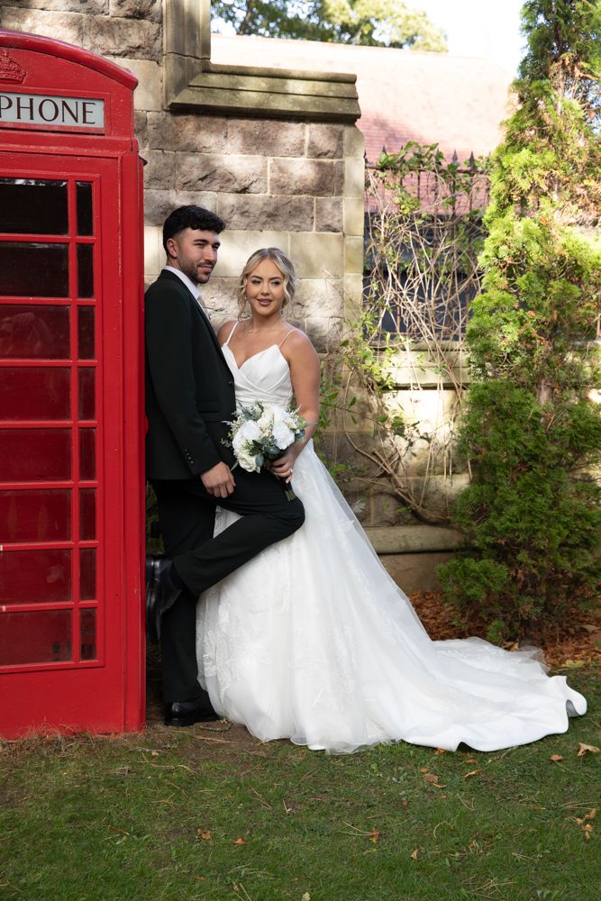 A romantic wedding photo series featuring a couple posing by a classic British red telephone booth in a garden setting.