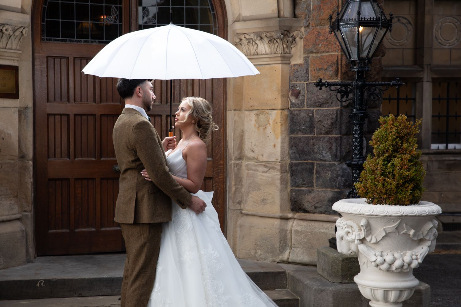 A bride and groom share an intimate moment under a white umbrella against a stone building backdrop.