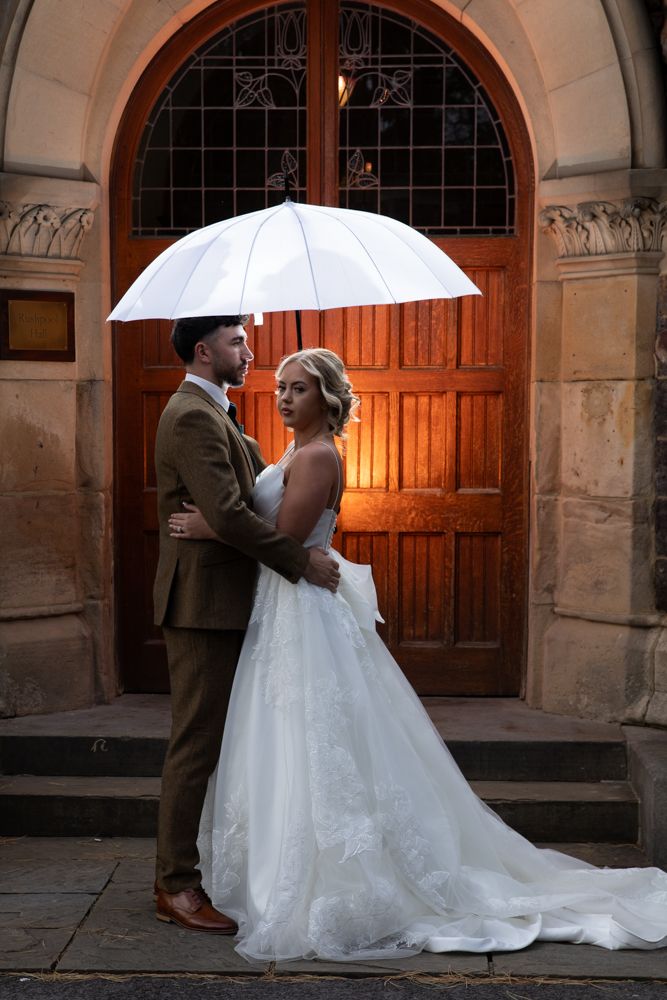 Bride and groom share romantic moments under an umbrella in an elegant stone archway at sunset.