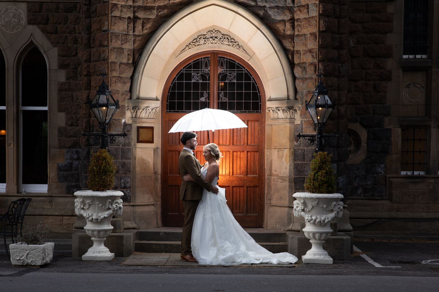 A wedding couple poses for photographs in front of a historic stone building with arched windows and ornate architecture.