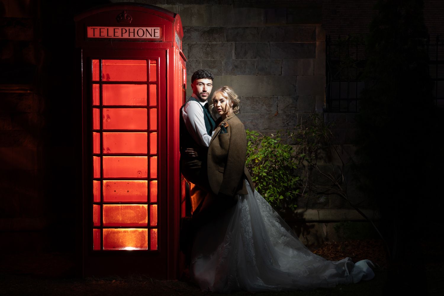A couple embraces near illuminated red British phone booths at night creating a romantic wedding scene.