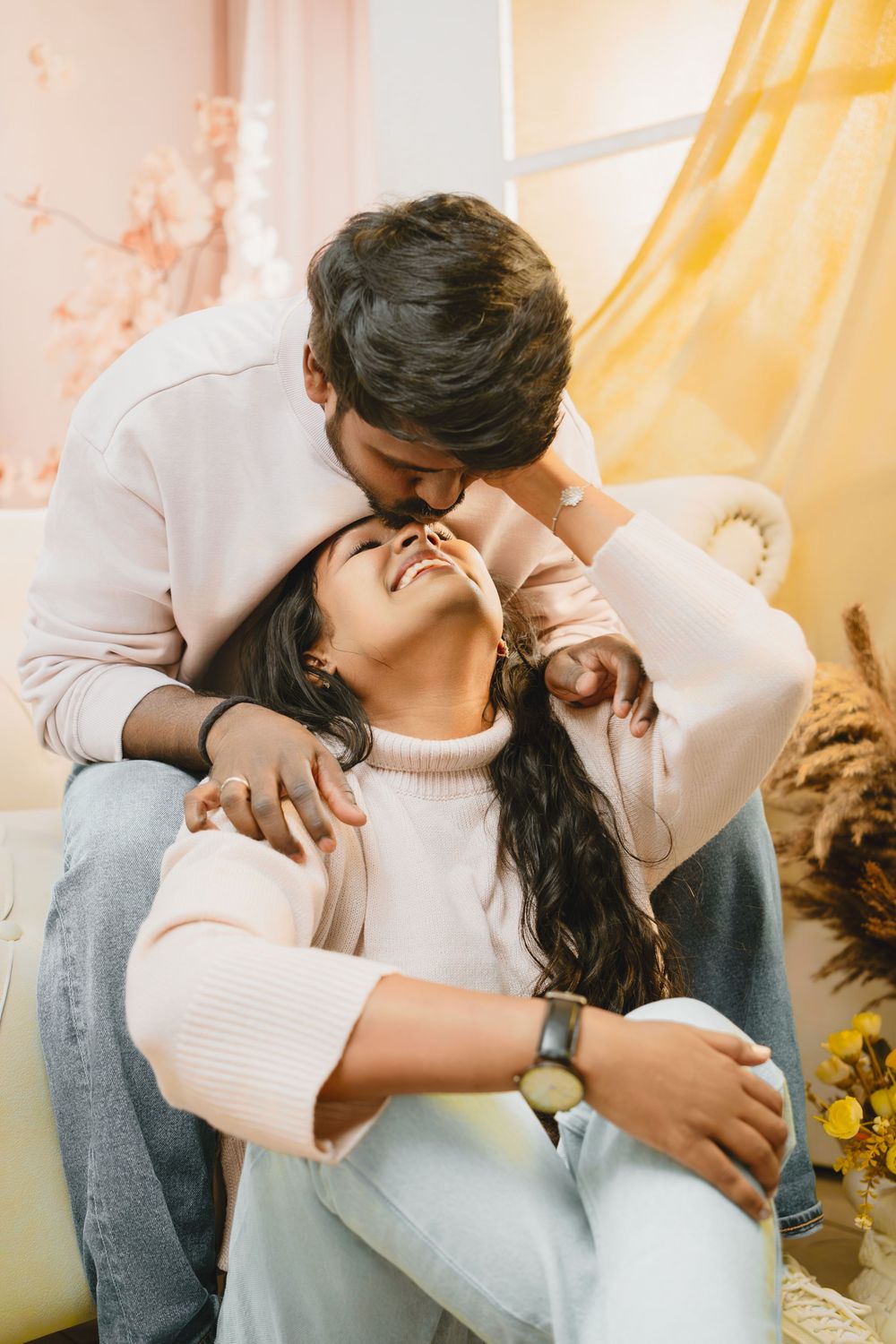 Romantic couple in white sweaters sharing an intimate moment near a sunny window with yellow curtains and sunflowers.