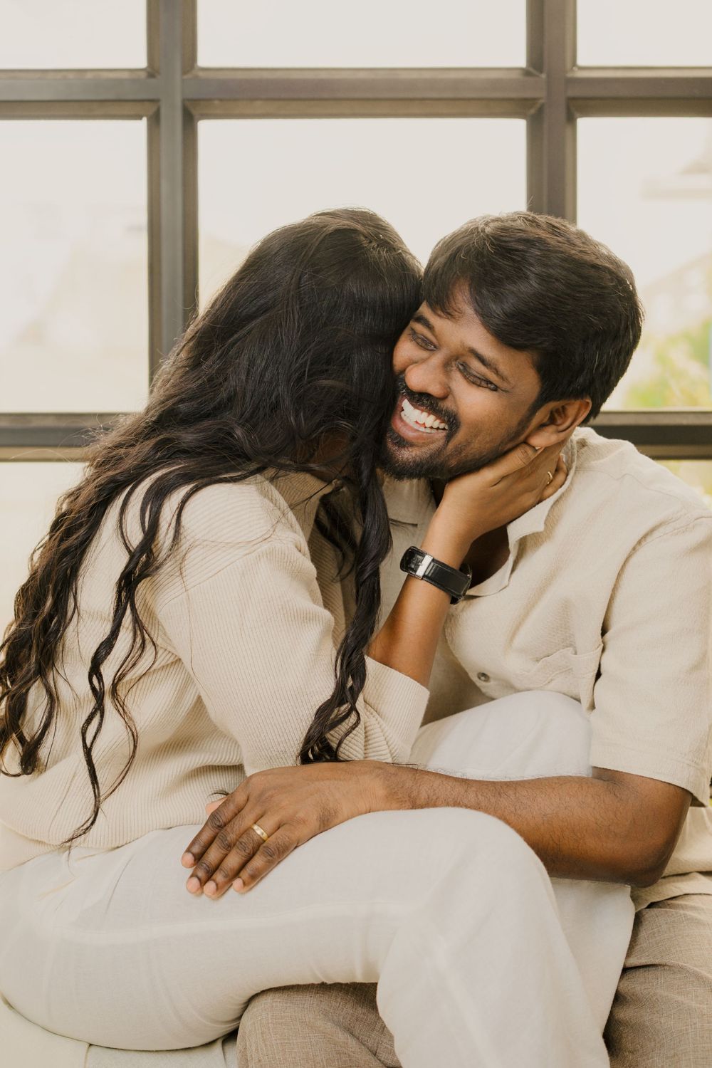 A couple in white outfits sharing intimate moments while sitting on a couch in a bright sunlit room.
