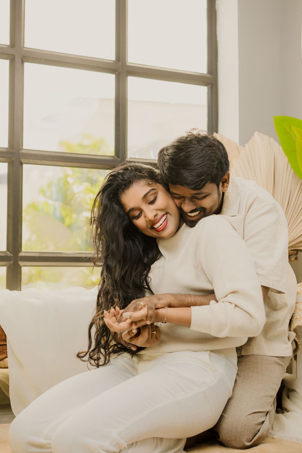 Couple in white outfits sharing intimate moments on a couch near bright windows in a living room.