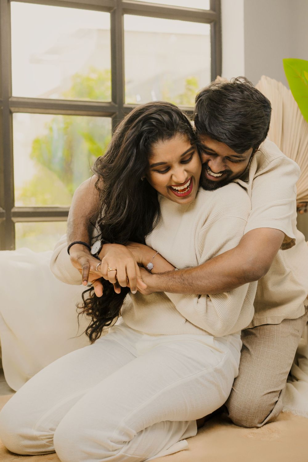 Couple dressed in cream colors share playful and affectionate moments on a couch by a window.