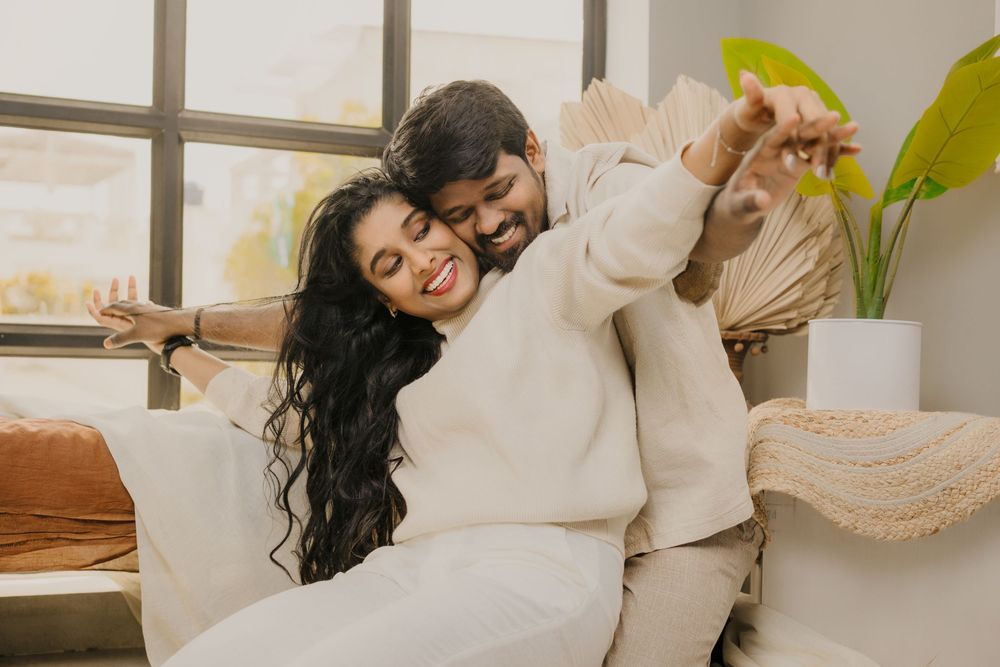Couple in cozy cream sweaters sharing a playful moment on white couch in bright living room with indoor plants.