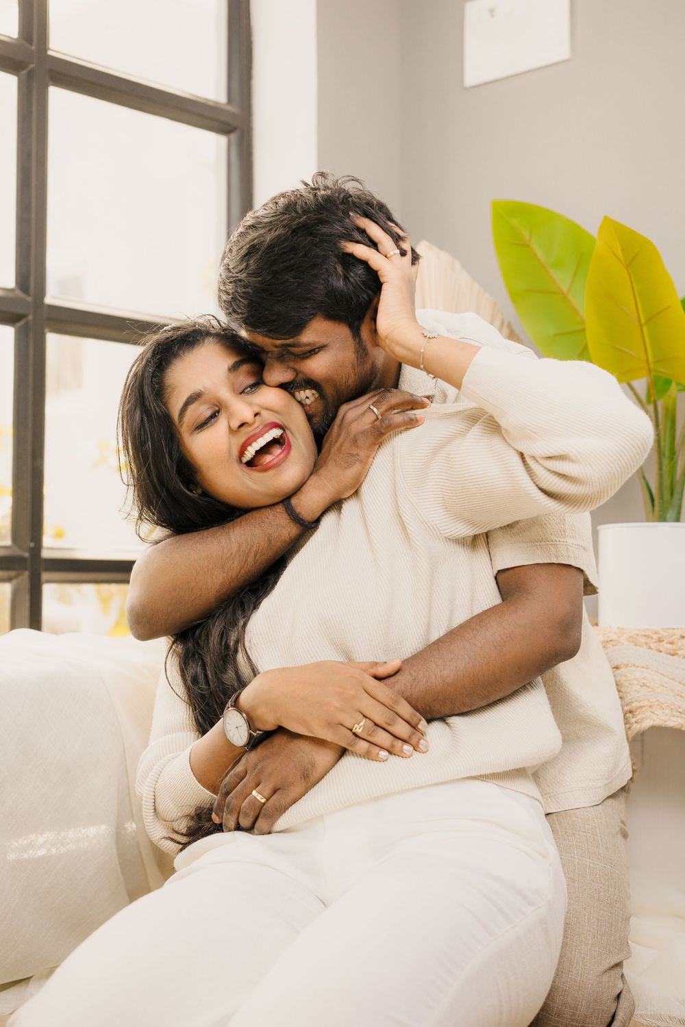 Romantic couple in white clothing sharing intimate moments and laughter by a bright window.