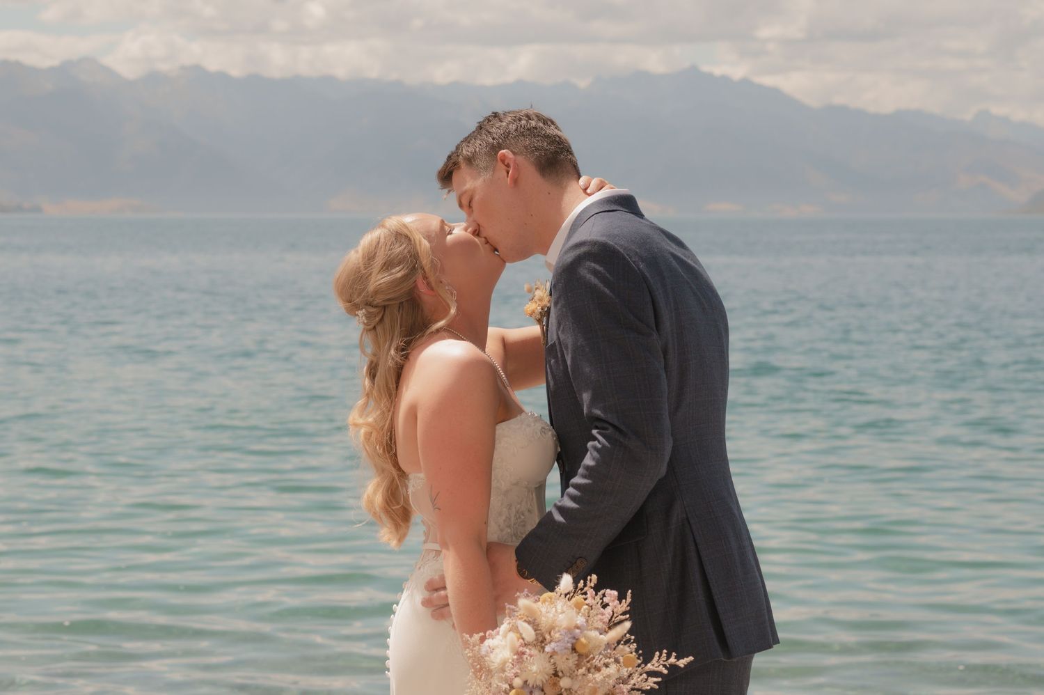 A couple shares a romantic kiss on a beach with mountains and turquoise waters in the background.