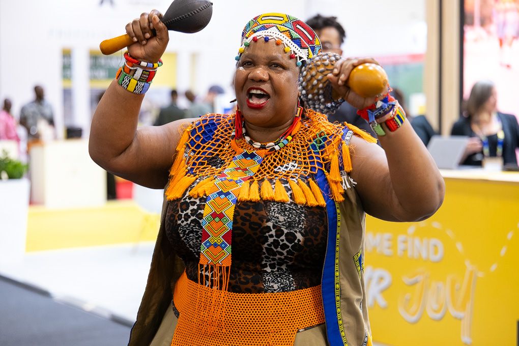 A South African performer in vibrant orange traditional dress and beaded necklace shakes maracas during a performance at the 2025 WTM.