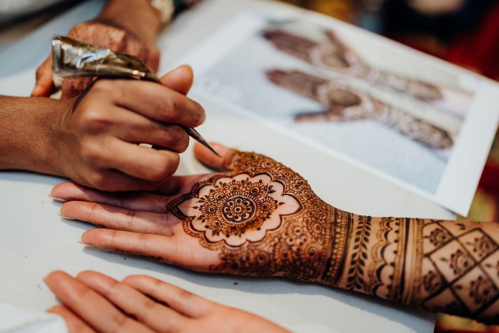Close-up of intricate henna design being applied to a hand with reference book in background.