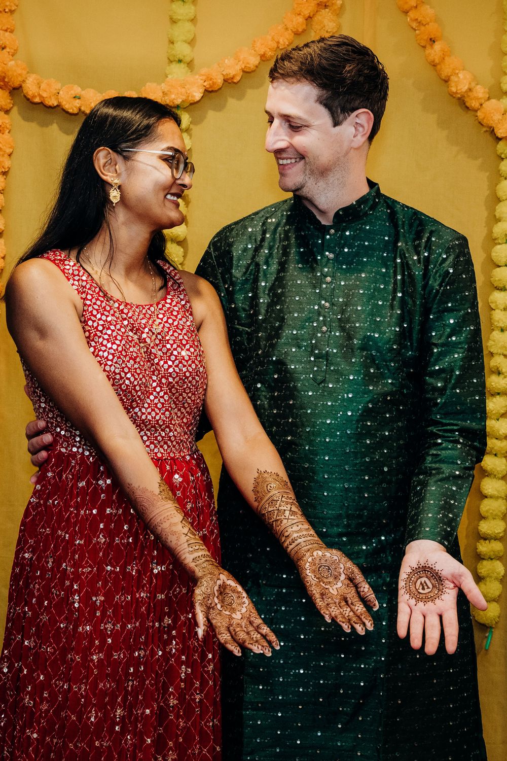 Couple in traditional red and green attire showing off mehndi designs against yellow backdrop.