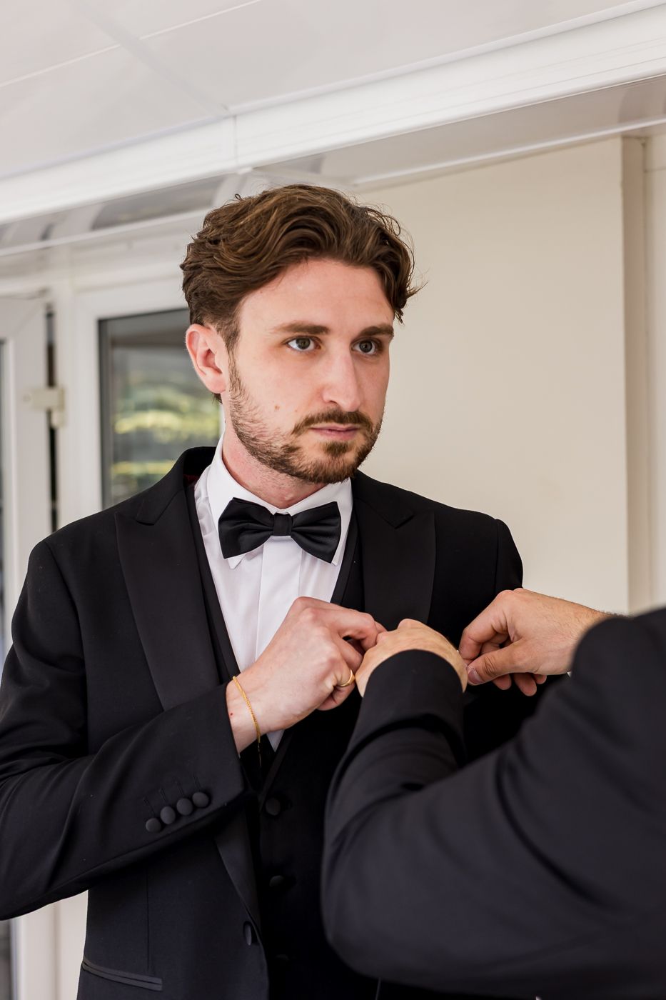 Formal wedding attire being adjusted with black tuxedo and bow tie in bright indoor setting.