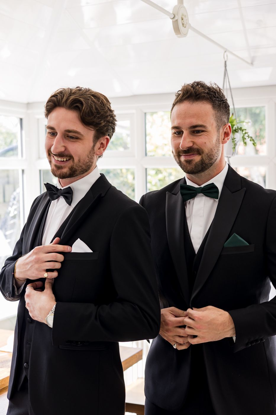 Two groomsmen in matching black tuxedos and bow ties stand together in a bright indoor space.