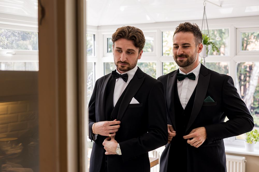 Two groomsmen in classic black tuxedos prepare for formal event in sunlit room.