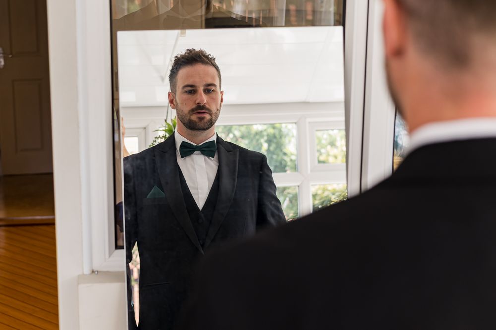 Groomsman in black tuxedo and bow tie looks in mirror while getting ready.