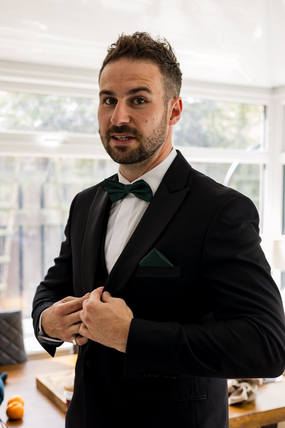 Groomsman in black tuxedo with emerald green pocket square adjusts his formal attire.