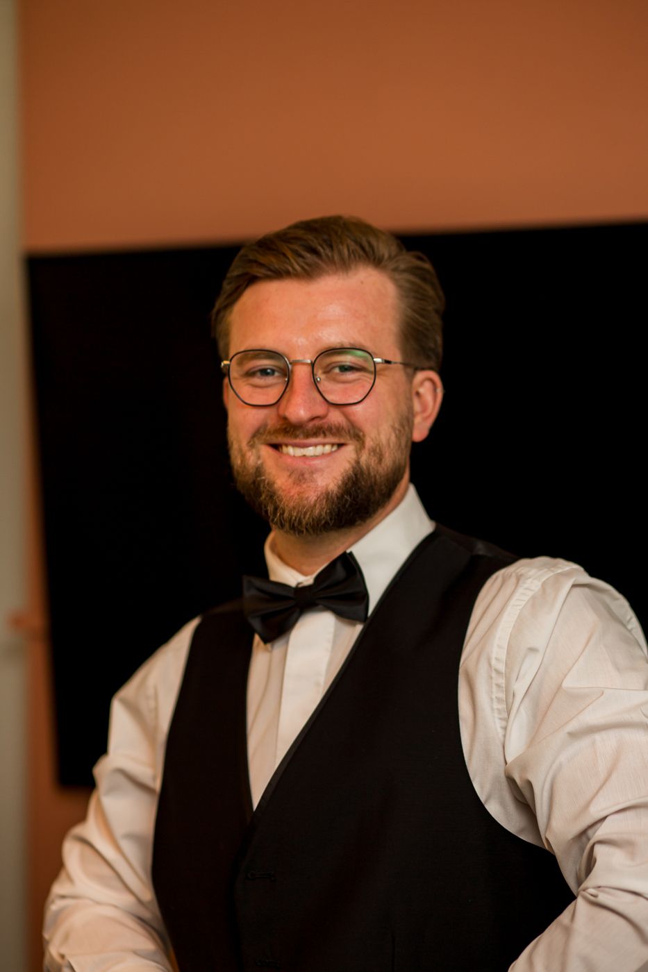 Groomsman in vest and bow tie smiles warmly against orange wall background.