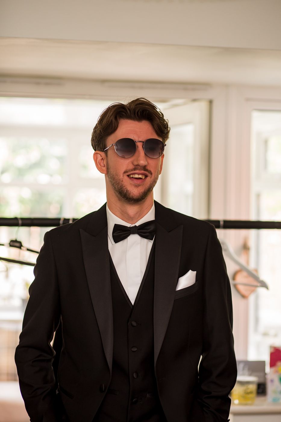 Stylish groomsman in black tuxedo and sunglasses smiles in bright indoor setting.