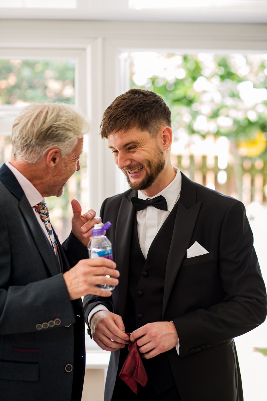 Groomsmen in black tuxedos share a lighthearted moment while getting ready for a wedding celebration.