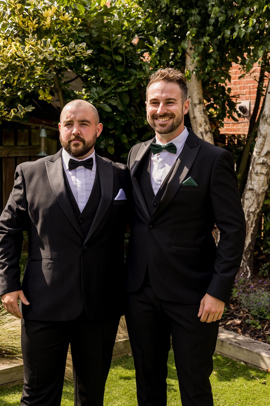 Wedding party members in formal black suits pose together outdoors with trees in the background.