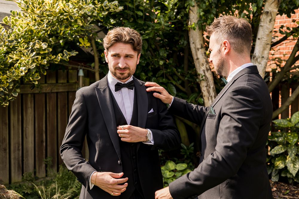 Two groomsmen in formal attire help each other prepare outdoors in garden setting.