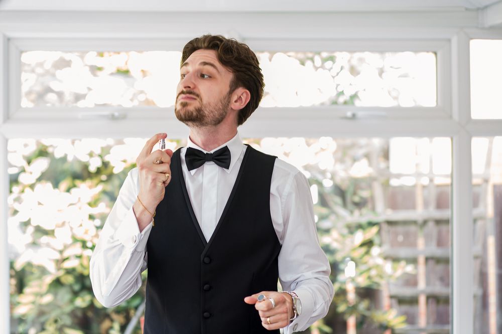 Groomsman adjusts his bow tie while wearing a formal black vest and white dress shirt in a sunlit room.