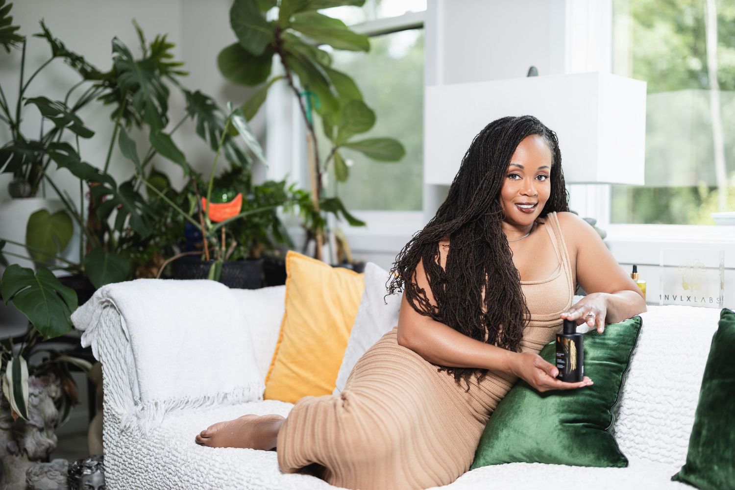 A person relaxes on a white couch with green throw pillows surrounded by lush indoor plants.