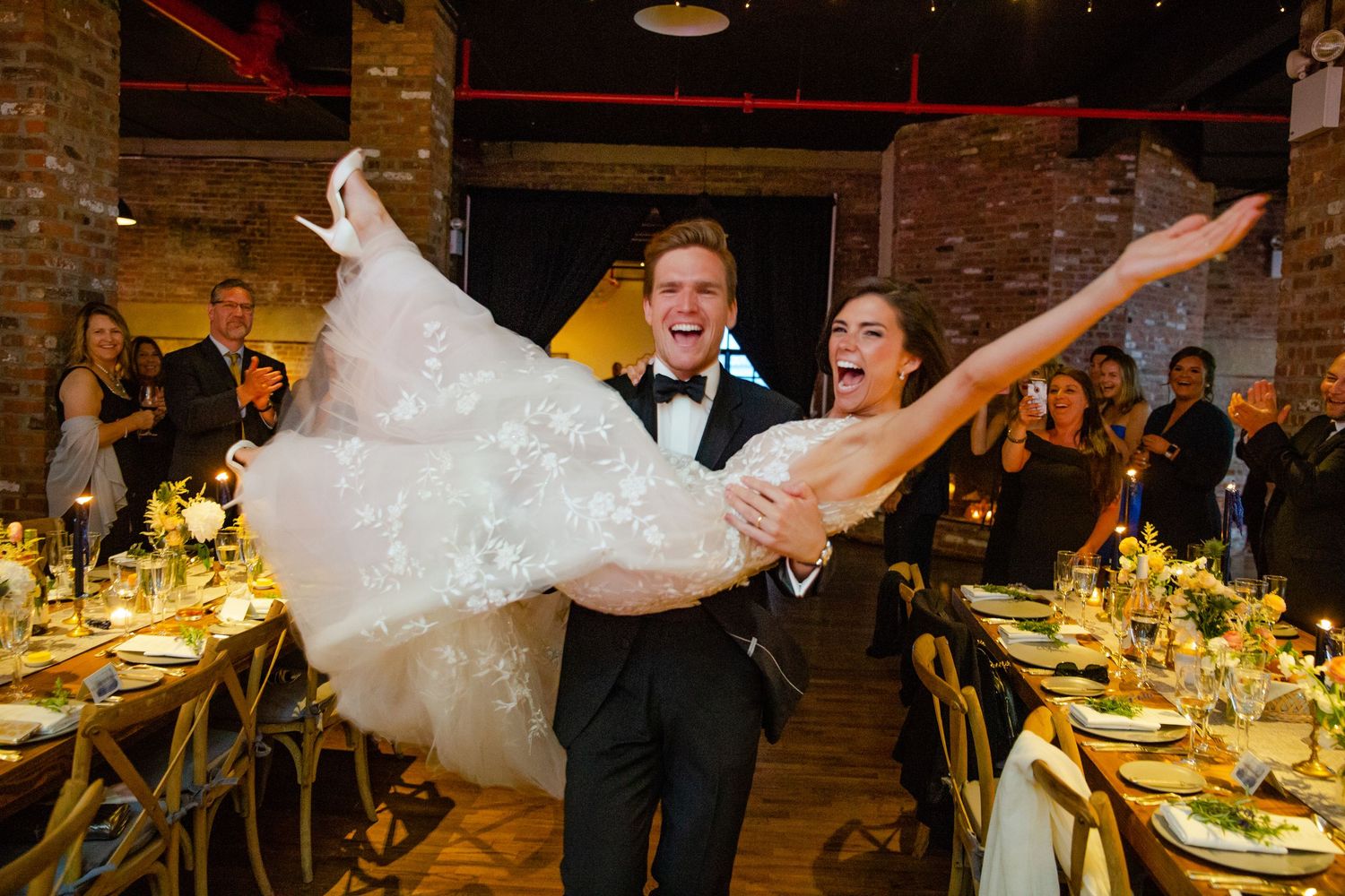 Wedding reception guests celebrate at elegantly decorated tables under twinkling lights in a brick-walled venue.