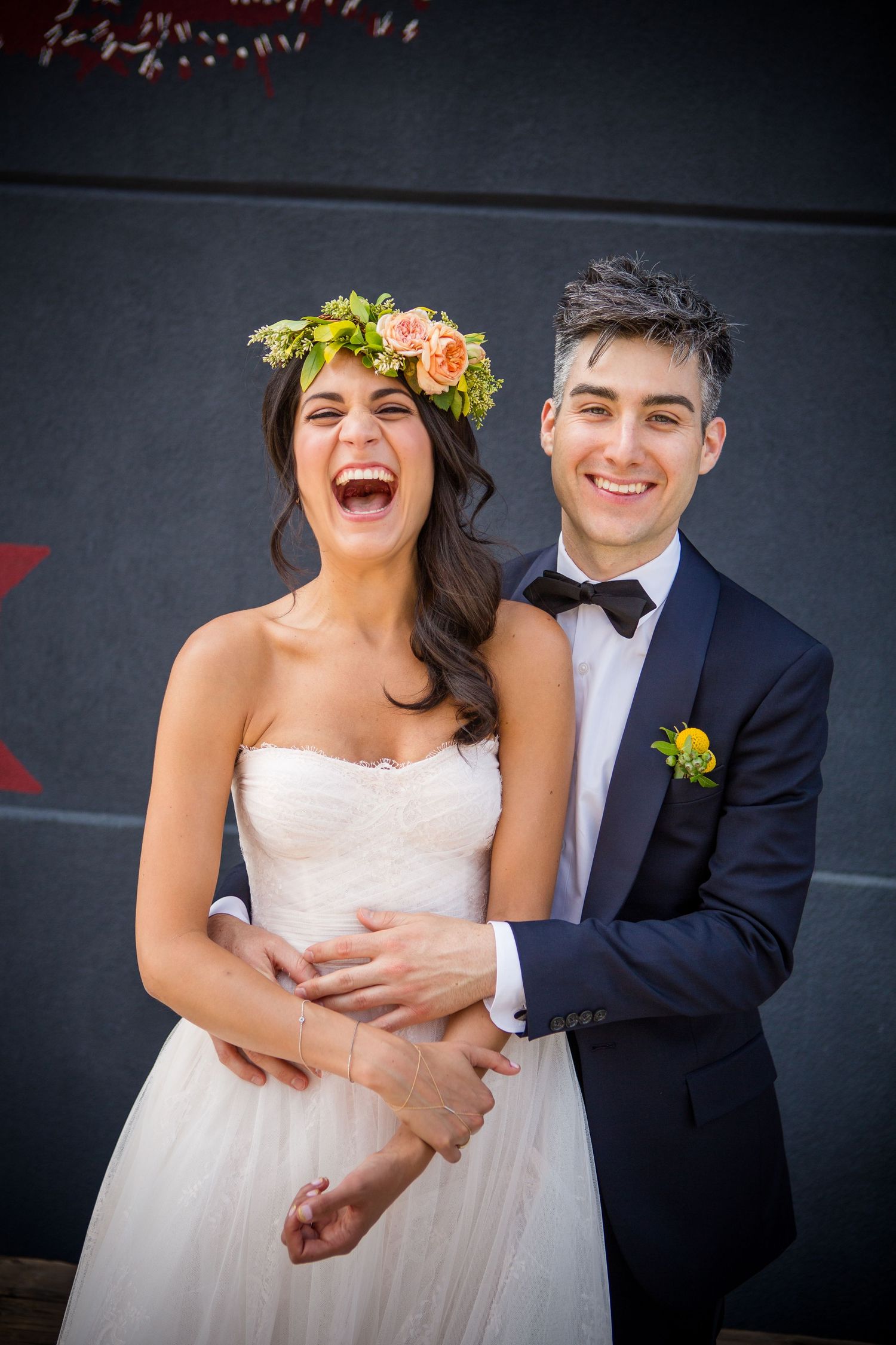 A joyful wedding couple shares candid laughter together against a dark wall during their photo session.