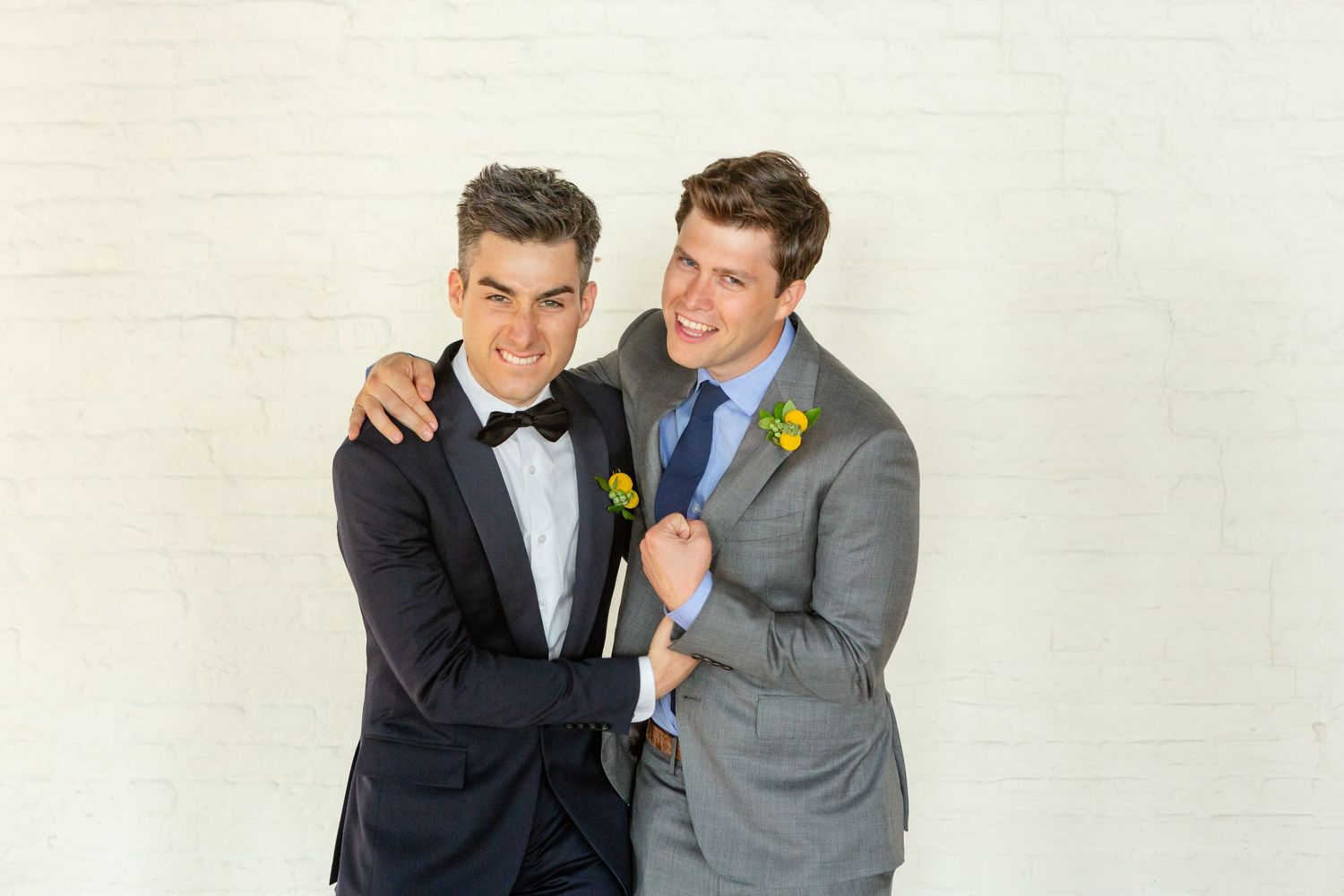 Two people in formal attire share a happy embrace while wearing boutonnieres against a light background.