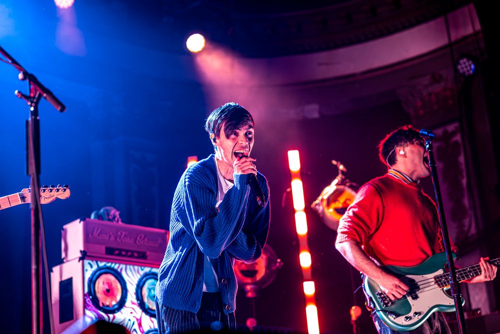Musicians performing on stage with colorful purple and blue lighting during a live rock concert performance.