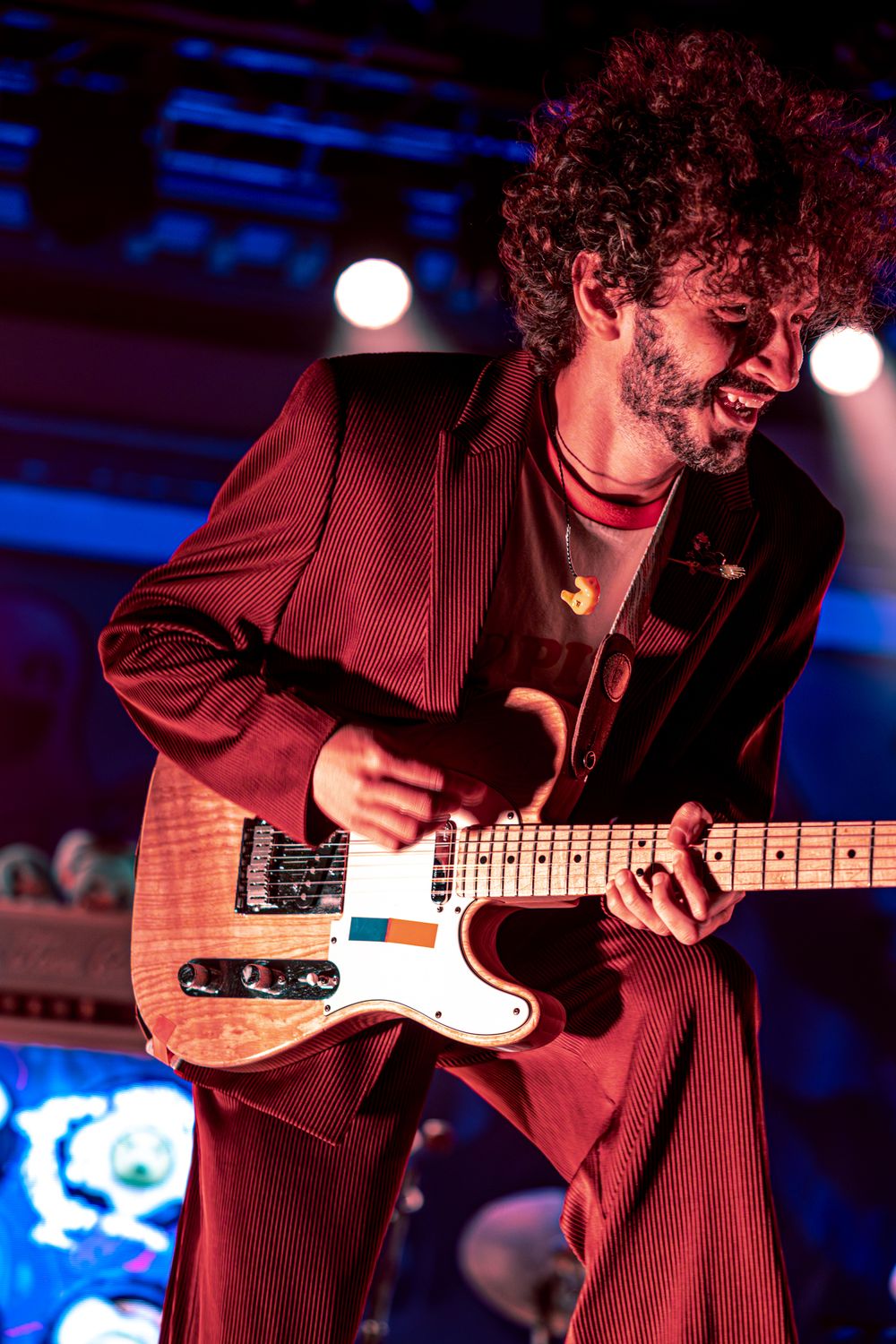 Guitarist in red lighting performs on stage with a Telecaster-style electric guitar against blue background lighting.