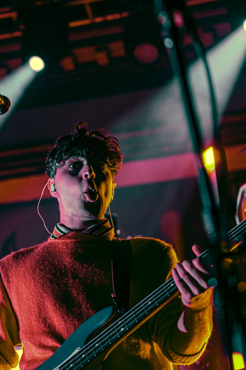 Musician plays bass guitar in red sweater under dramatic concert lighting with colorful stage effects.