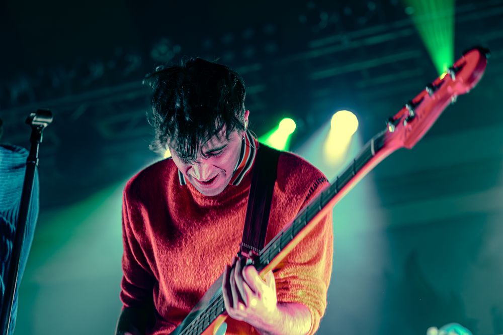 A guitarist performs on stage bathed in vibrant green and blue lighting effects during a live concert performance.