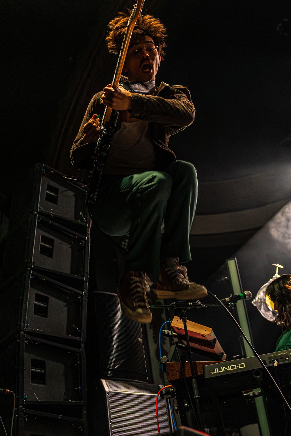 Musician plays electric bass guitar on dark stage with dramatic lighting and speaker stacks visible in background.