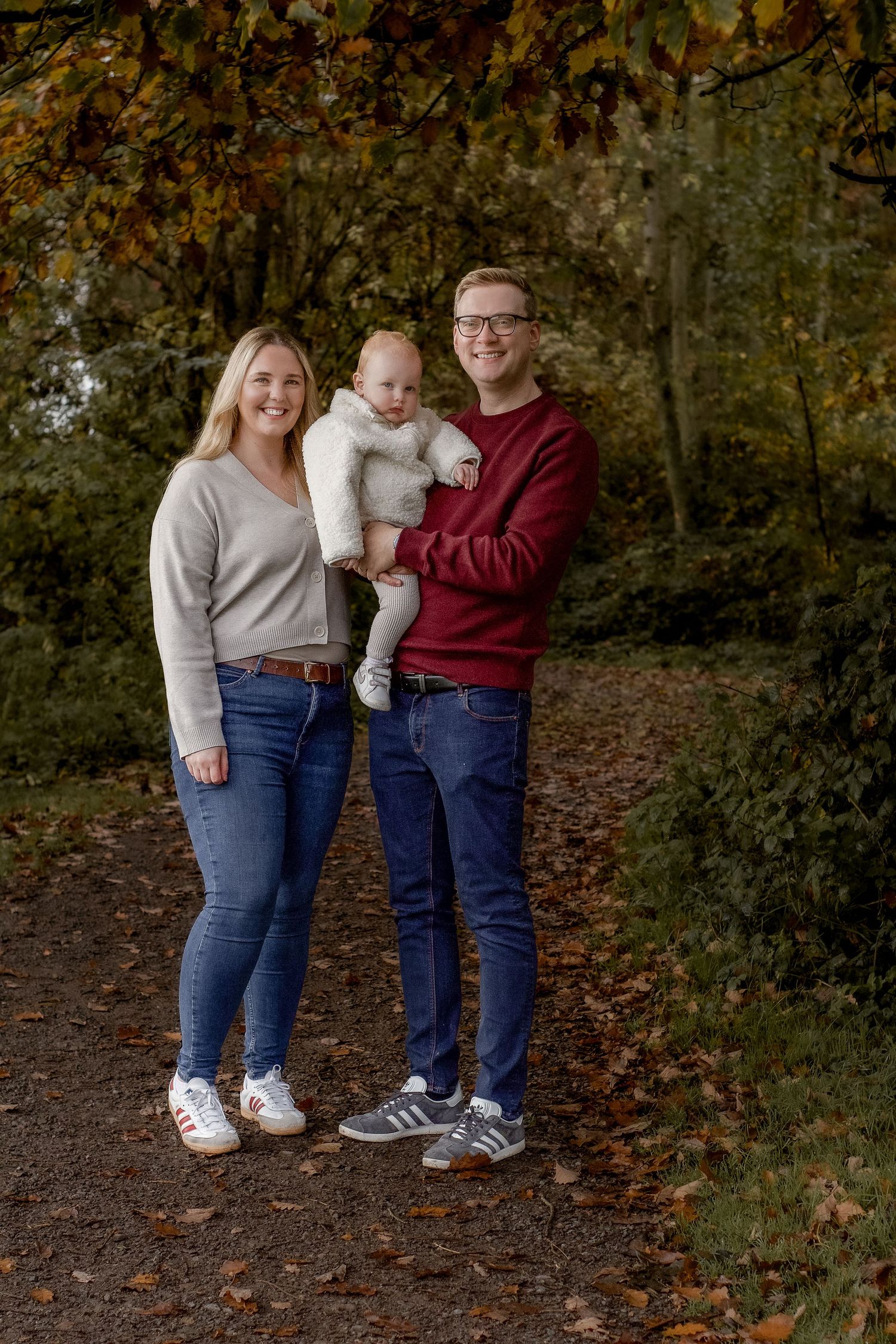 Family wearing coordinating outfits of burgundy and beige stands together on autumn path covered in fallen leaves.
