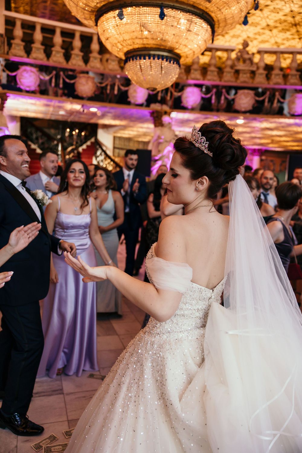 A bride in a sparkling white gown dances with guests under ornate chandeliers at an elegant ballroom reception.