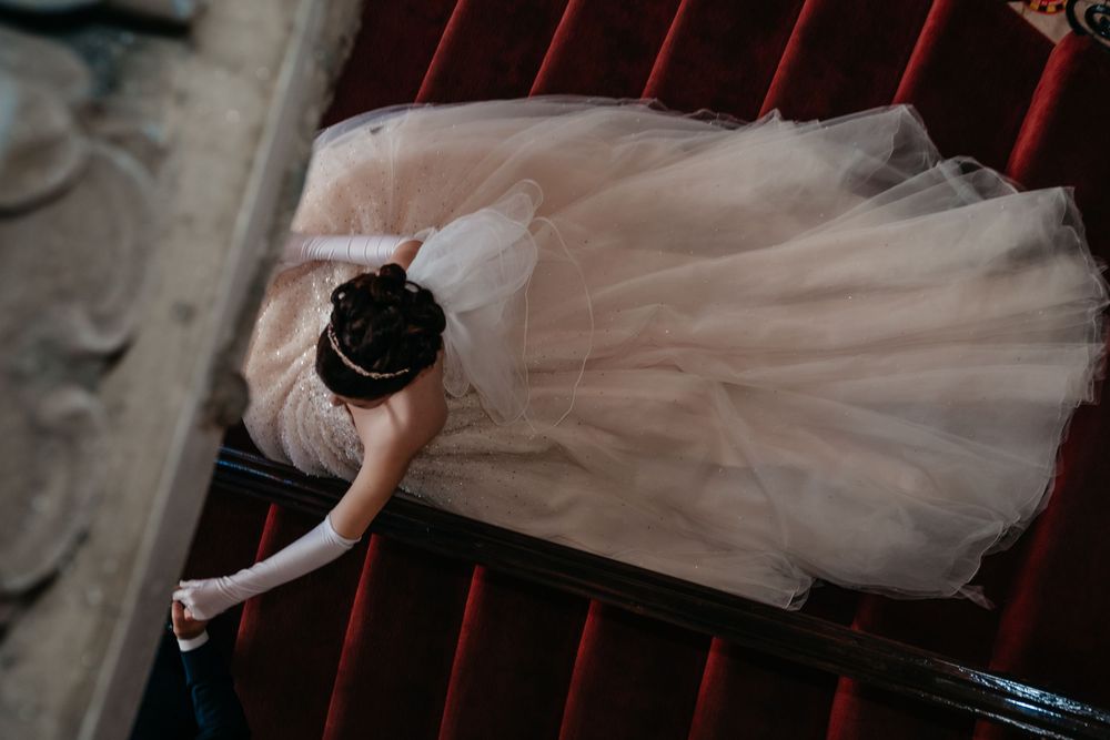 A bride in a flowing white dress walks down a dark staircase with dramatic lighting and shadows.
