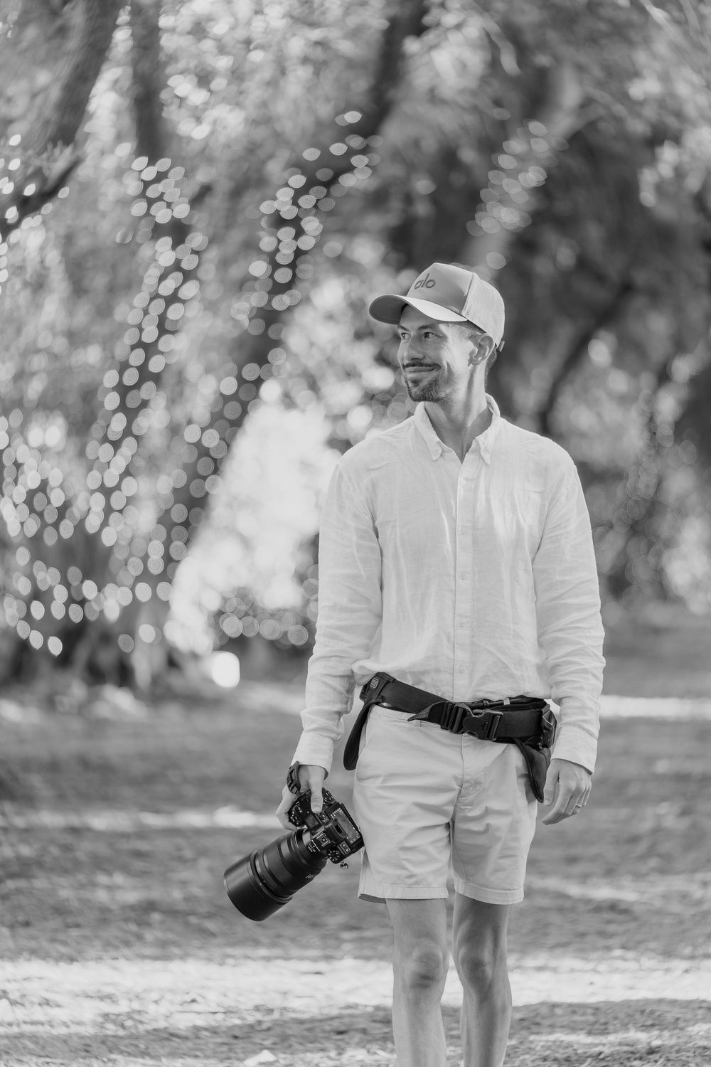 A photographer in casual white attire and baseball cap holds a professional camera while walking outdoors in a dreamy setting.