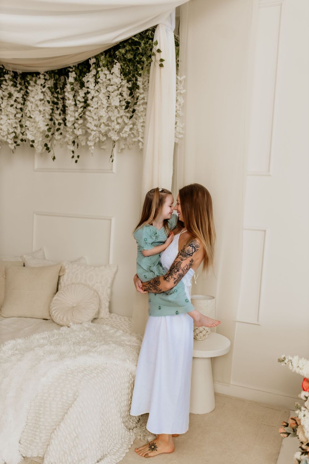 A tattooed mother holds her young daughter in a bright, white bedroom decorated with hanging flowers.
