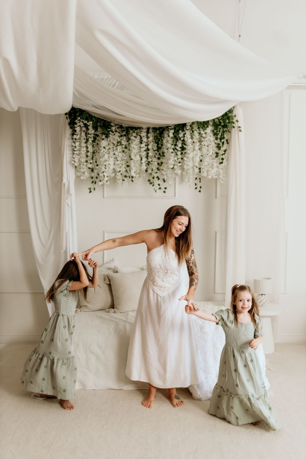 A woman in a white dress dances with two girls in sage green dresses in an elegant floral bedroom.