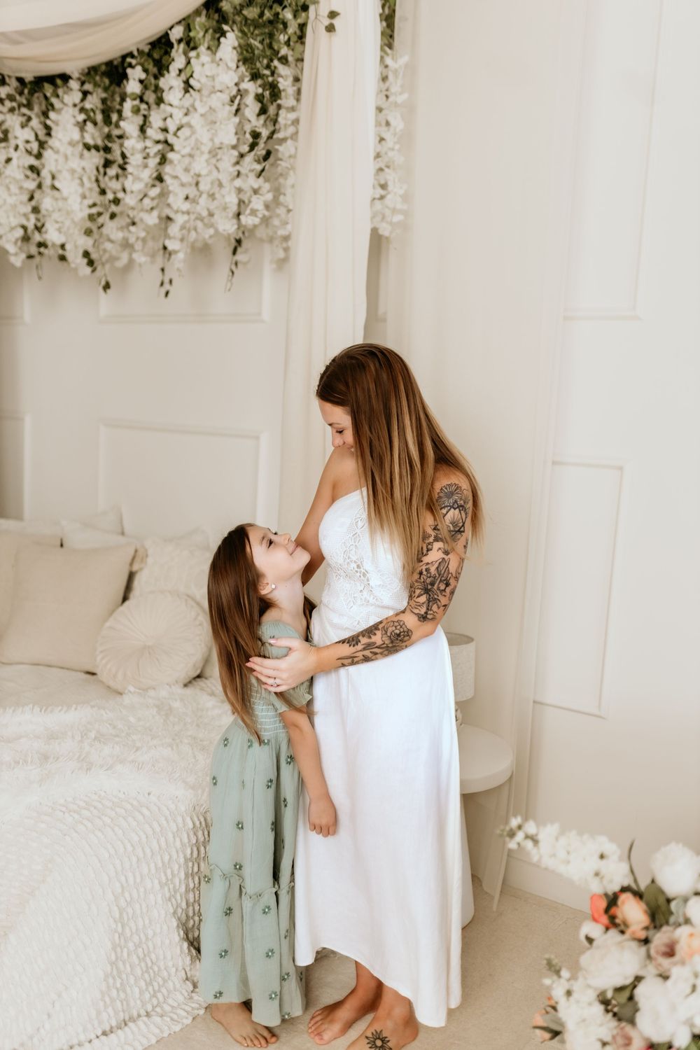 A tattooed mother in a white dress embraces her young daughter in a floral-decorated white bedroom.