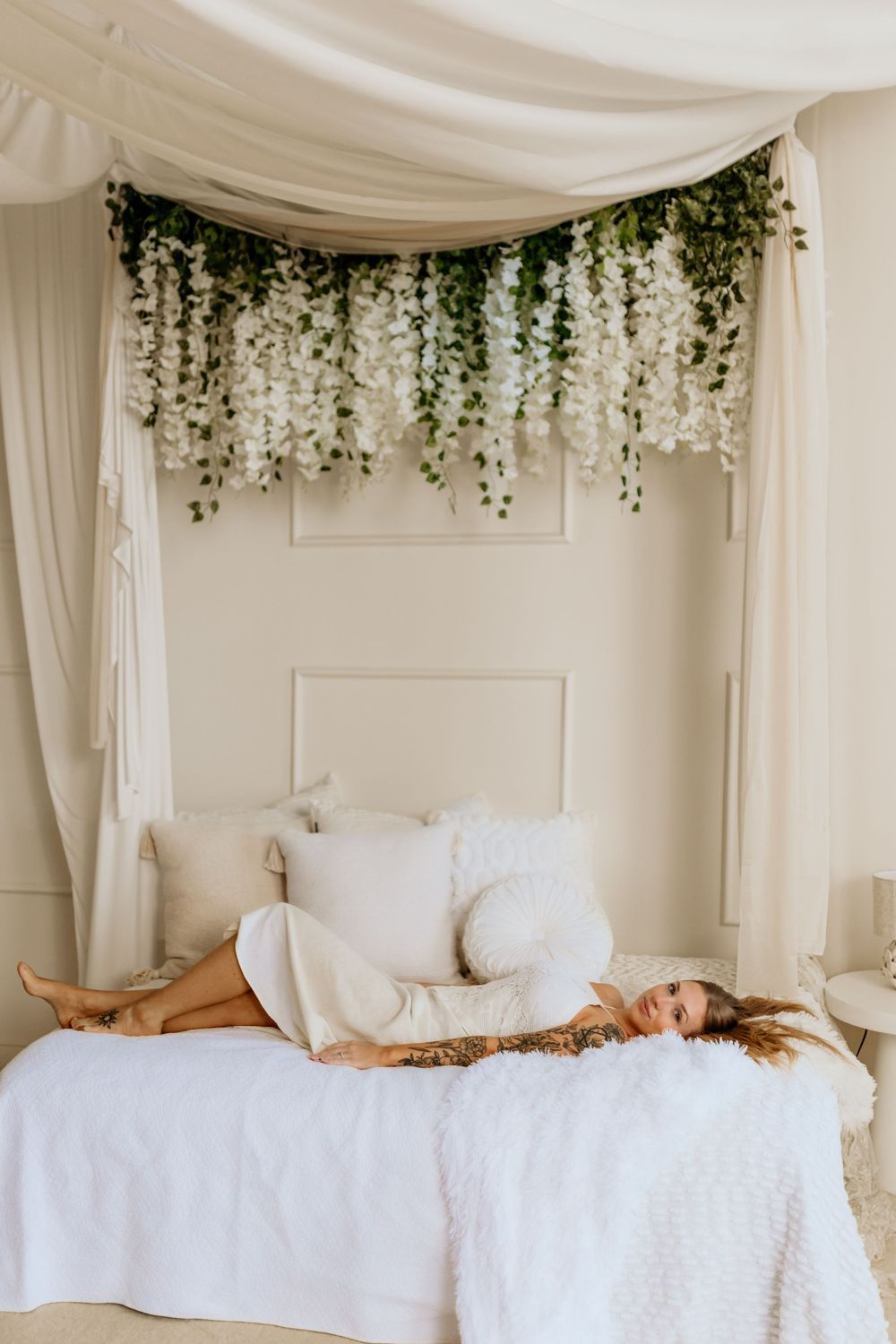 Woman in white outfit relaxing on a canopy bed adorned with cascading white flowers and greenery.