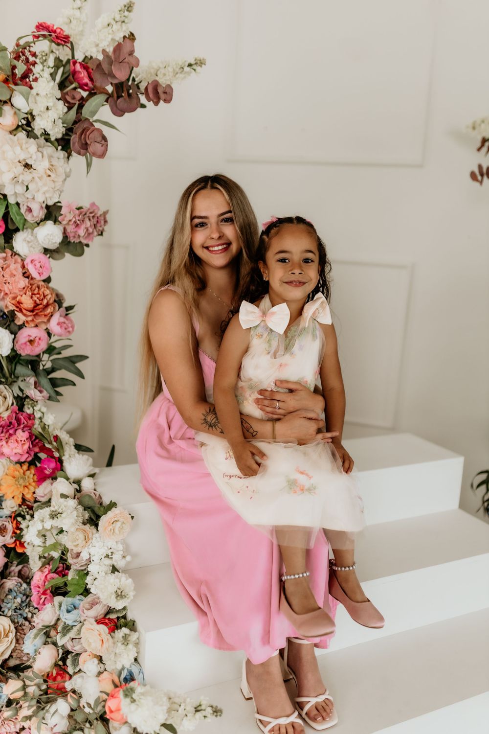 Woman in pink dress and young girl in floral dress pose together on white steps surrounded by colorful flowers.