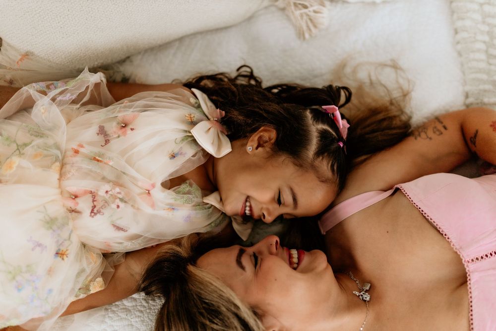 Two girls lying down laughing together, one in floral dress with white bow, other in pink top with pink bow.