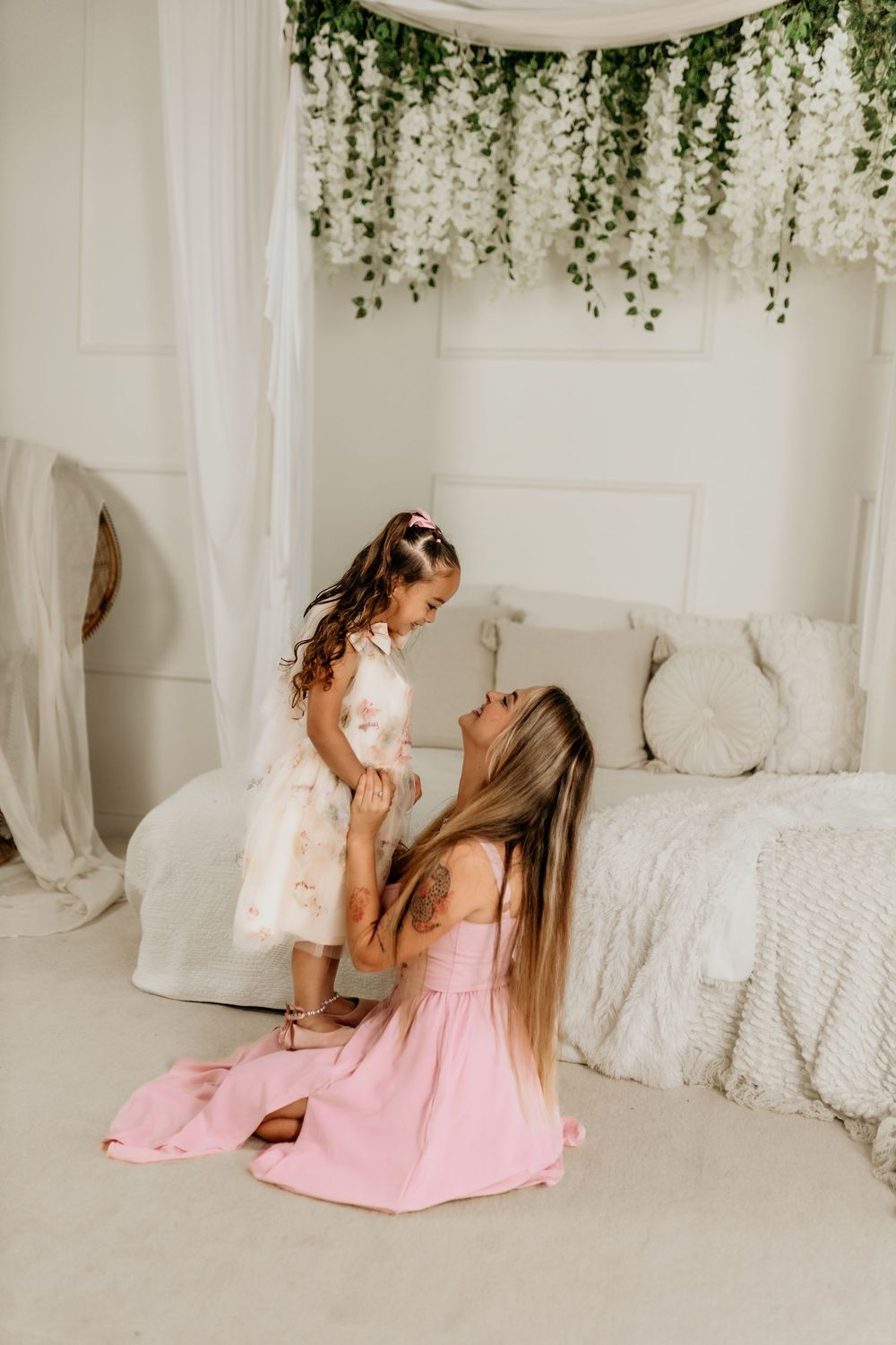 A mother in a pink dress kneels on the floor holding her young daughter in a floral dress in a white bedroom.